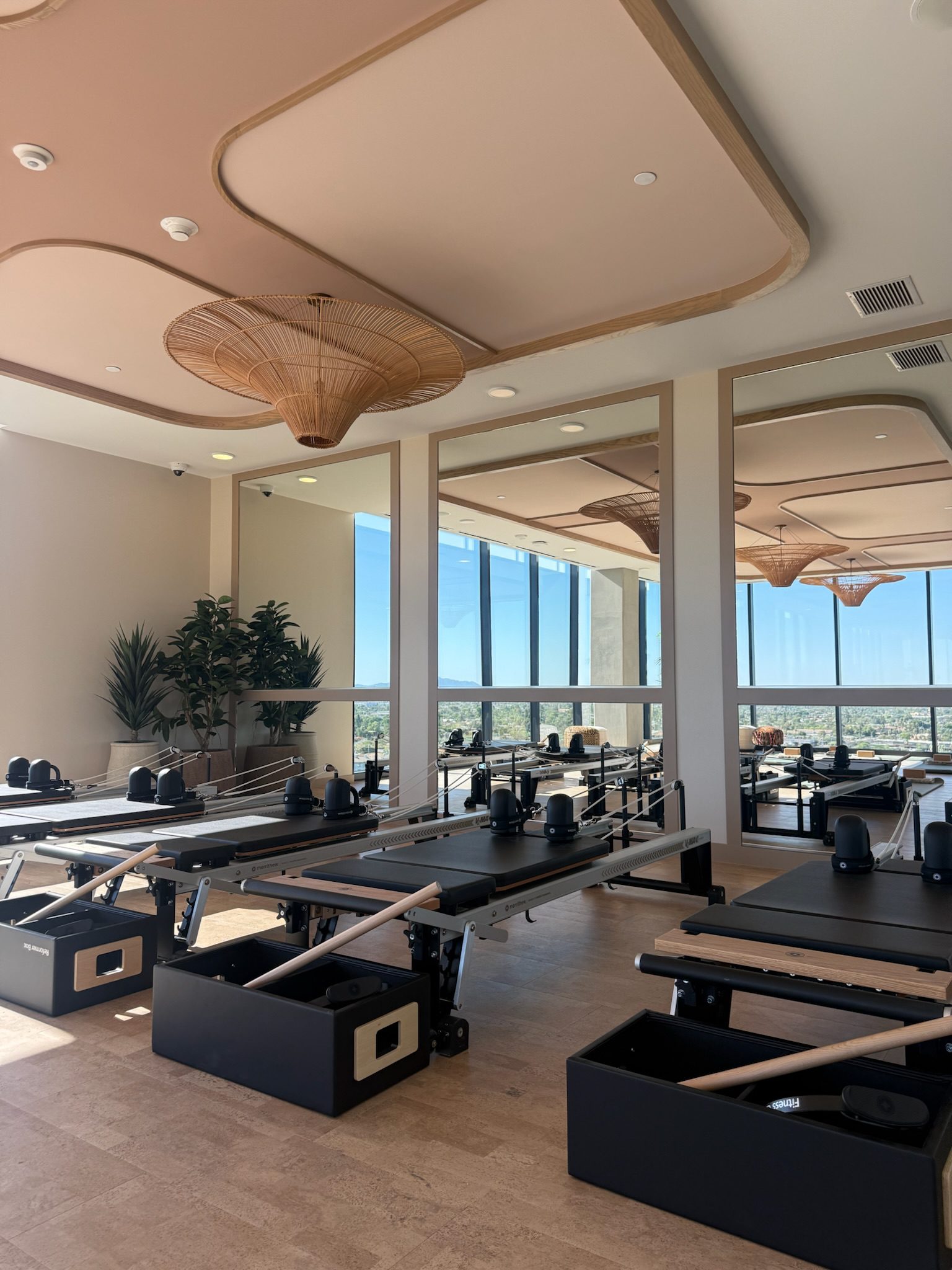 A bright, modern Pilates studio with several black reformer machines, large mirrors on the walls, tall windows letting in natural light, and a view of the city outside. Decorative ceiling fixtures and plants add to the ambiance.