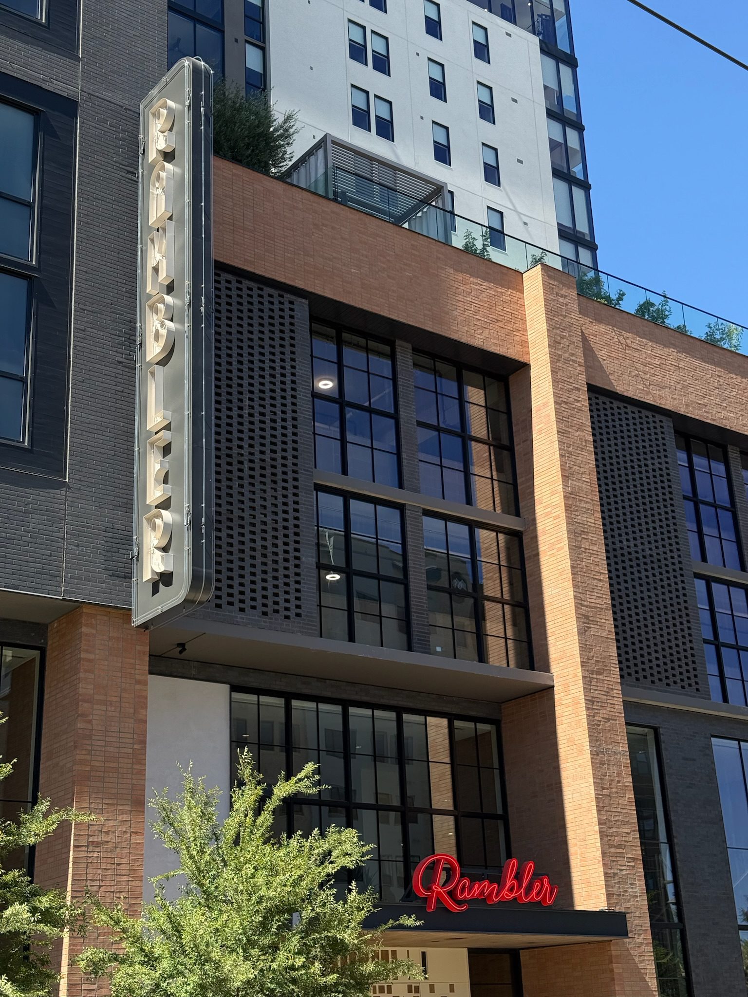 A modern brick and glass building with a vertical neon sign reading Rambler and a red neon Rambler sign above the entrance. Trees and a tall residential tower are also visible.
