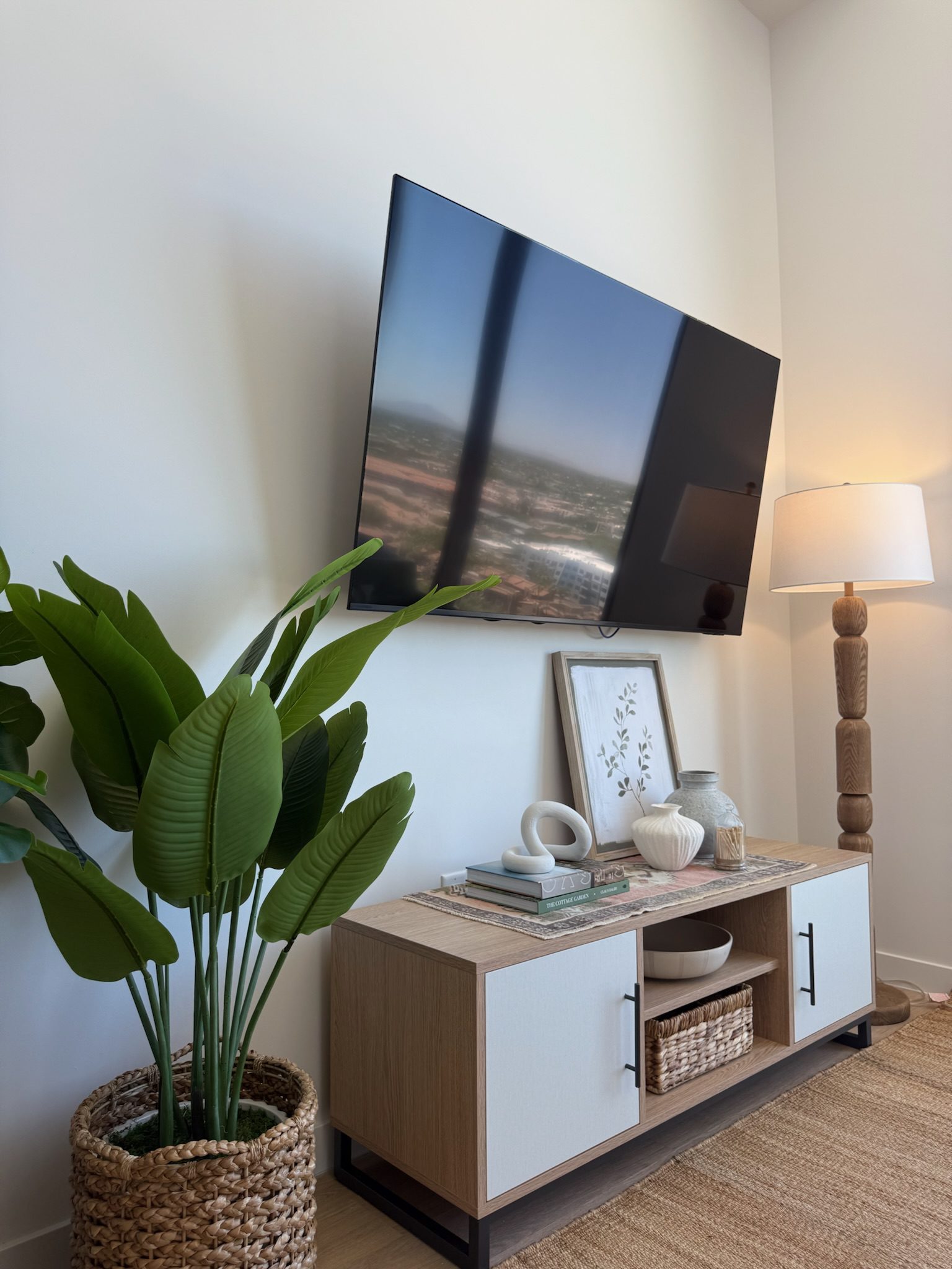 A modern living room corner with a wall-mounted TV, a potted plant, a wooden floor lamp, and a light wood console table decorated with art, ceramics, and a basket on the shelf below.