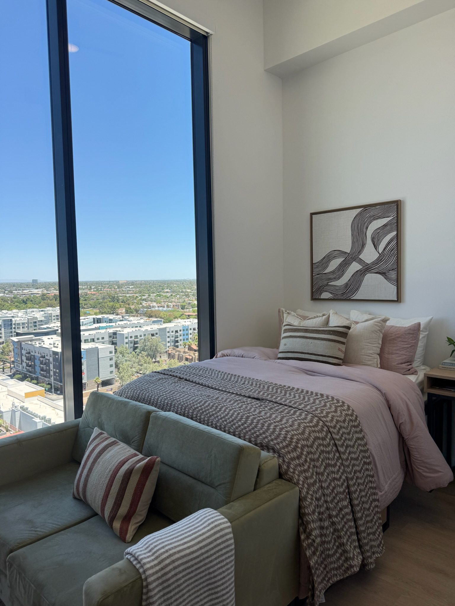 A cozy bedroom with a large window overlooking a cityscape. The bed has pink and gray bedding, pillows, and a patterned throw. A green sofa with striped pillows sits at the foot of the bed. Modern art hangs on the wall.