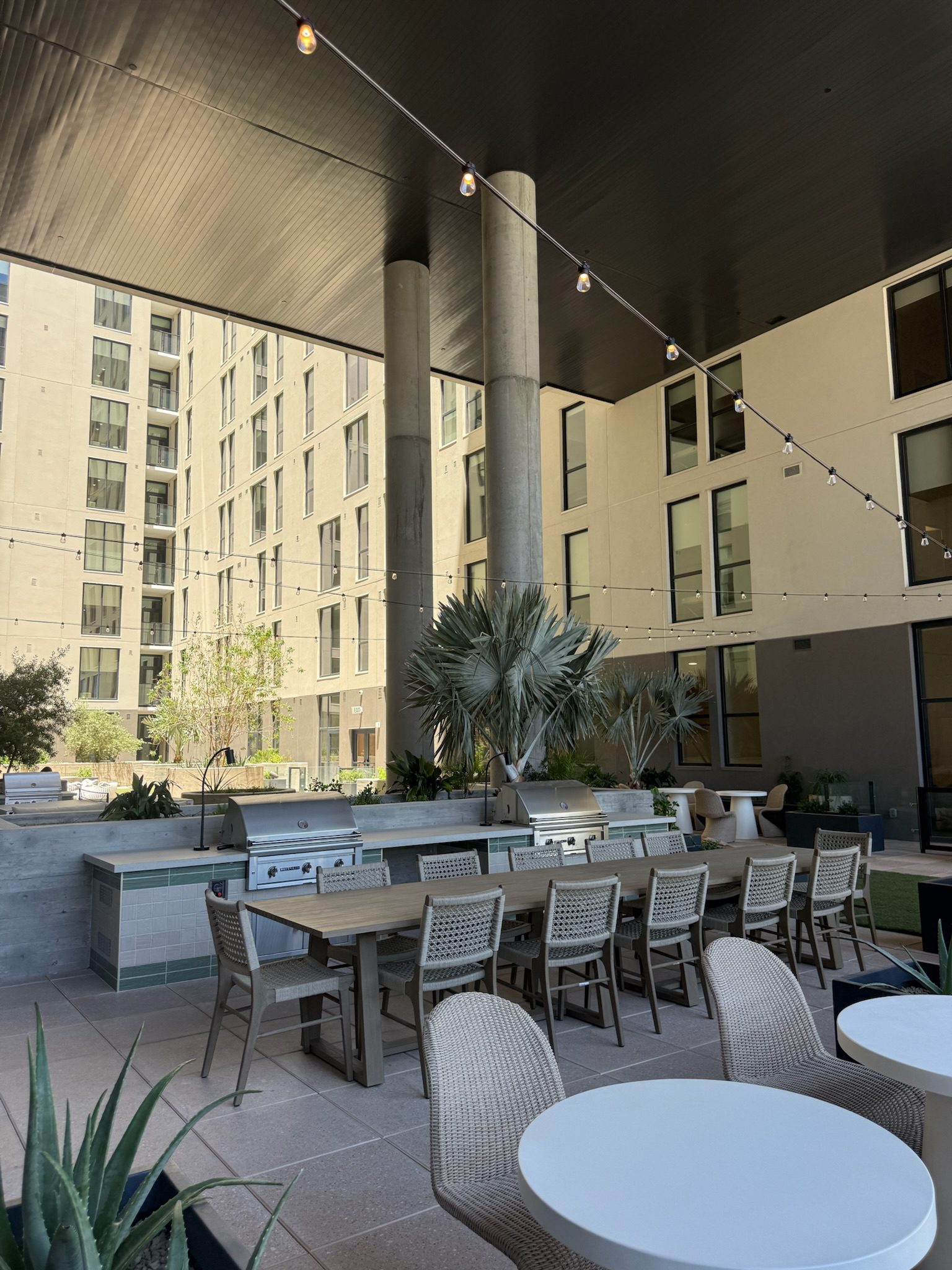 Outdoor communal dining area under a black canopy, featuring a long table with wicker chairs, built-in grills, string lights, potted plants, and surrounded by modern apartment buildings.
