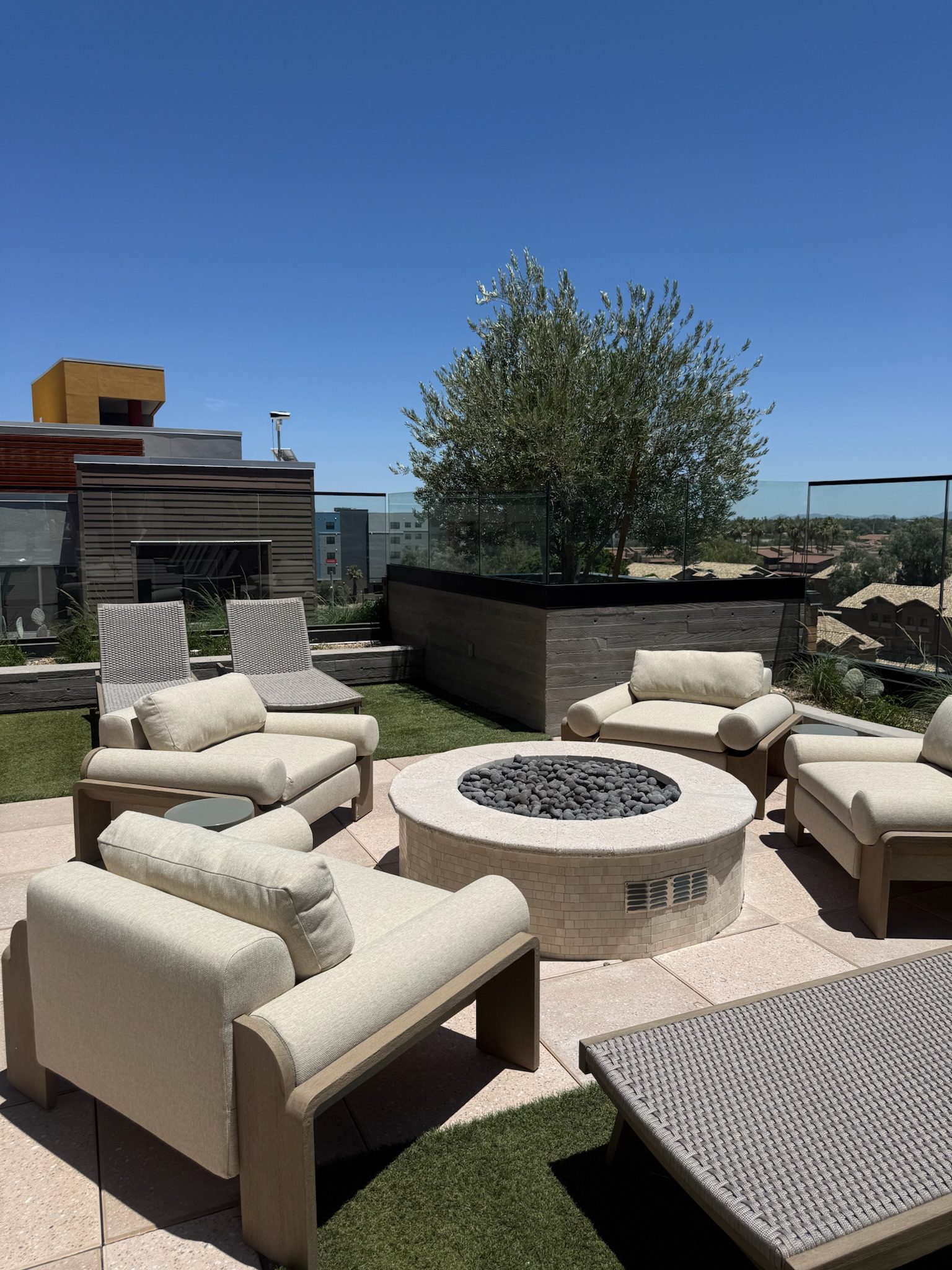 Modern rooftop patio with beige cushioned chairs arranged around a circular stone fire pit, artificial grass, a tree in a planter, and city buildings in the background under a clear blue sky.