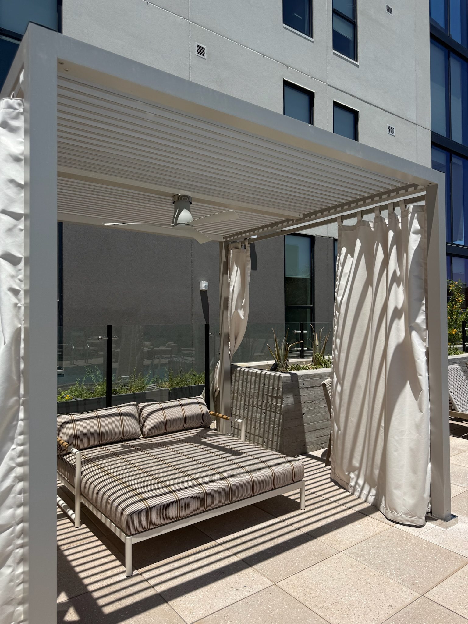 A modern outdoor cabana with white curtains, a striped cushioned daybed, and a slatted roof casts shadows on the patio, set against a tall building and some plants in the background.