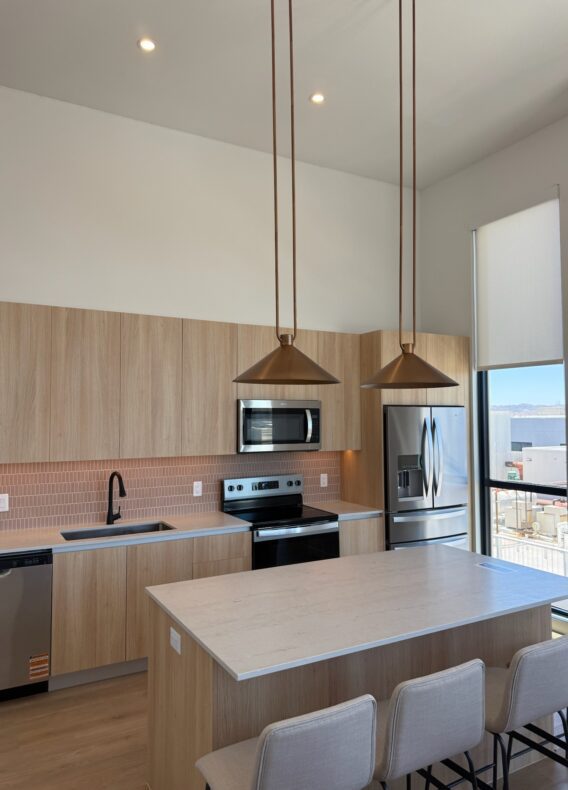 Modern kitchen with light wood cabinets, a large island with three upholstered stools, stainless steel appliances, two pendant lights, and a window with a view outside. Pink tile backsplash behind the sink and stove.