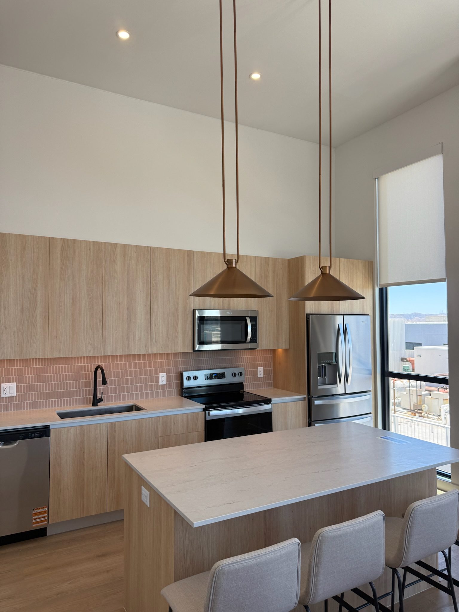 Modern kitchen with light wood cabinets, a large island with three upholstered stools, stainless steel appliances, two pendant lights, and a window with a view outside. Pink tile backsplash behind the sink and stove.