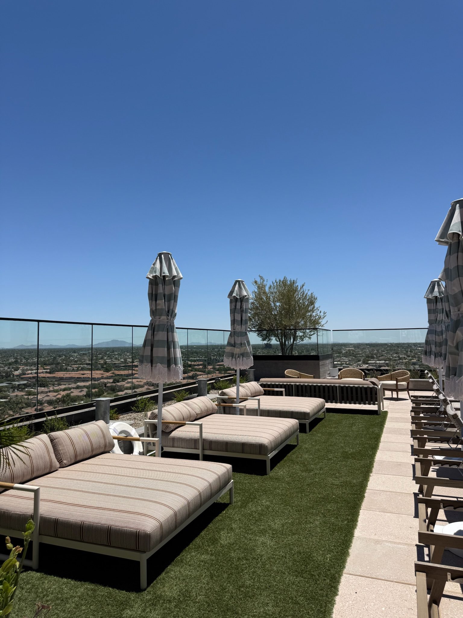 Rooftop lounge area with striped cushioned chairs, closed umbrellas, and glass railing overlooking a scenic city view under a clear blue sky.