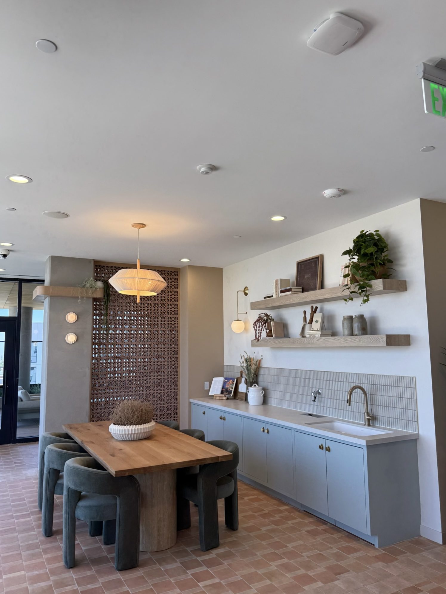 A modern kitchen and dining area with a wooden table, six green cushioned stools, light blue cabinets, open shelves with decor, a white tiled backsplash, and potted plants. The space is well-lit with natural and ceiling lights.