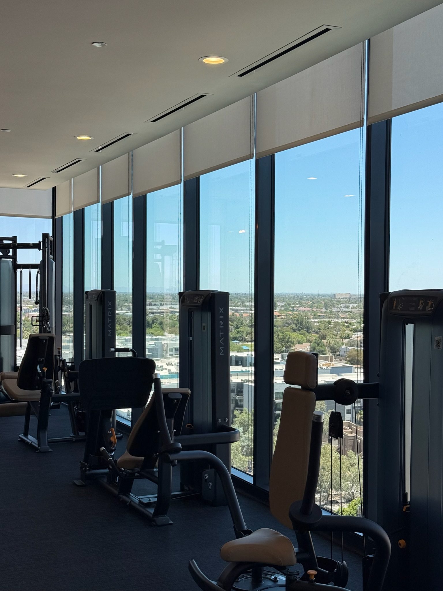A modern gym with exercise machines is situated by large floor-to-ceiling windows, offering a bright view of the cityscape and blue sky outside.