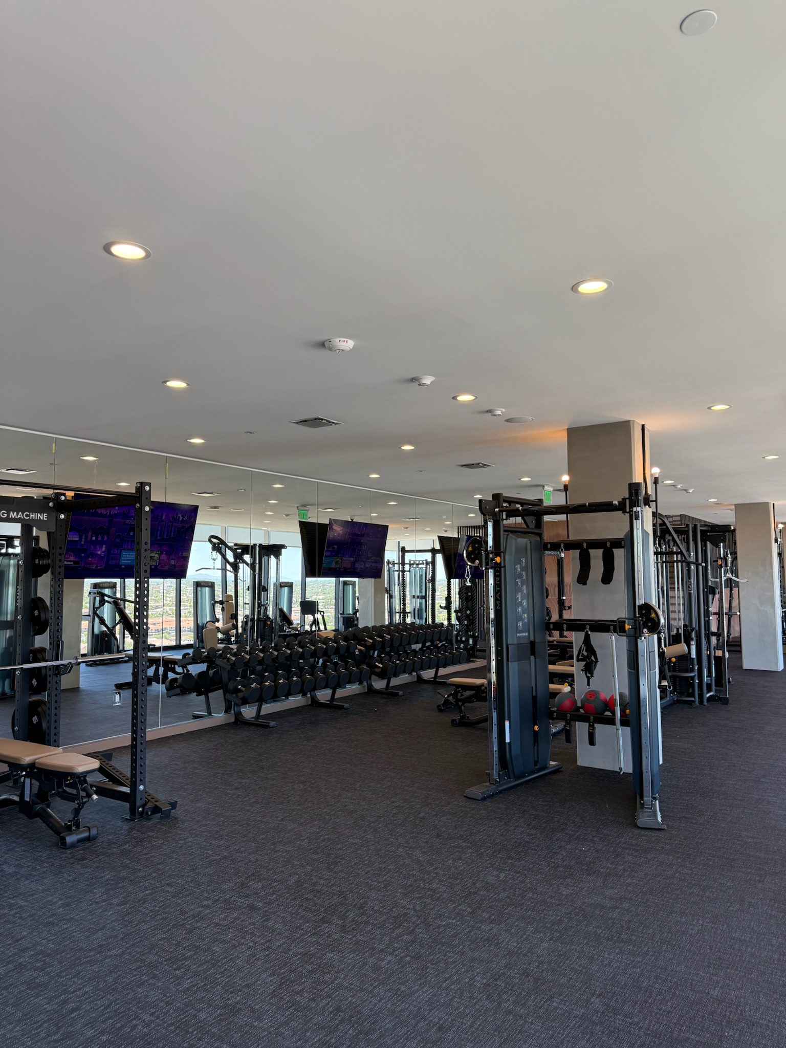 A modern gym with workout benches, dumbbells, weight racks, and cable machines. The room has large mirrors, ceiling lights, and two wall-mounted TVs, with natural light coming from windows on the right side.