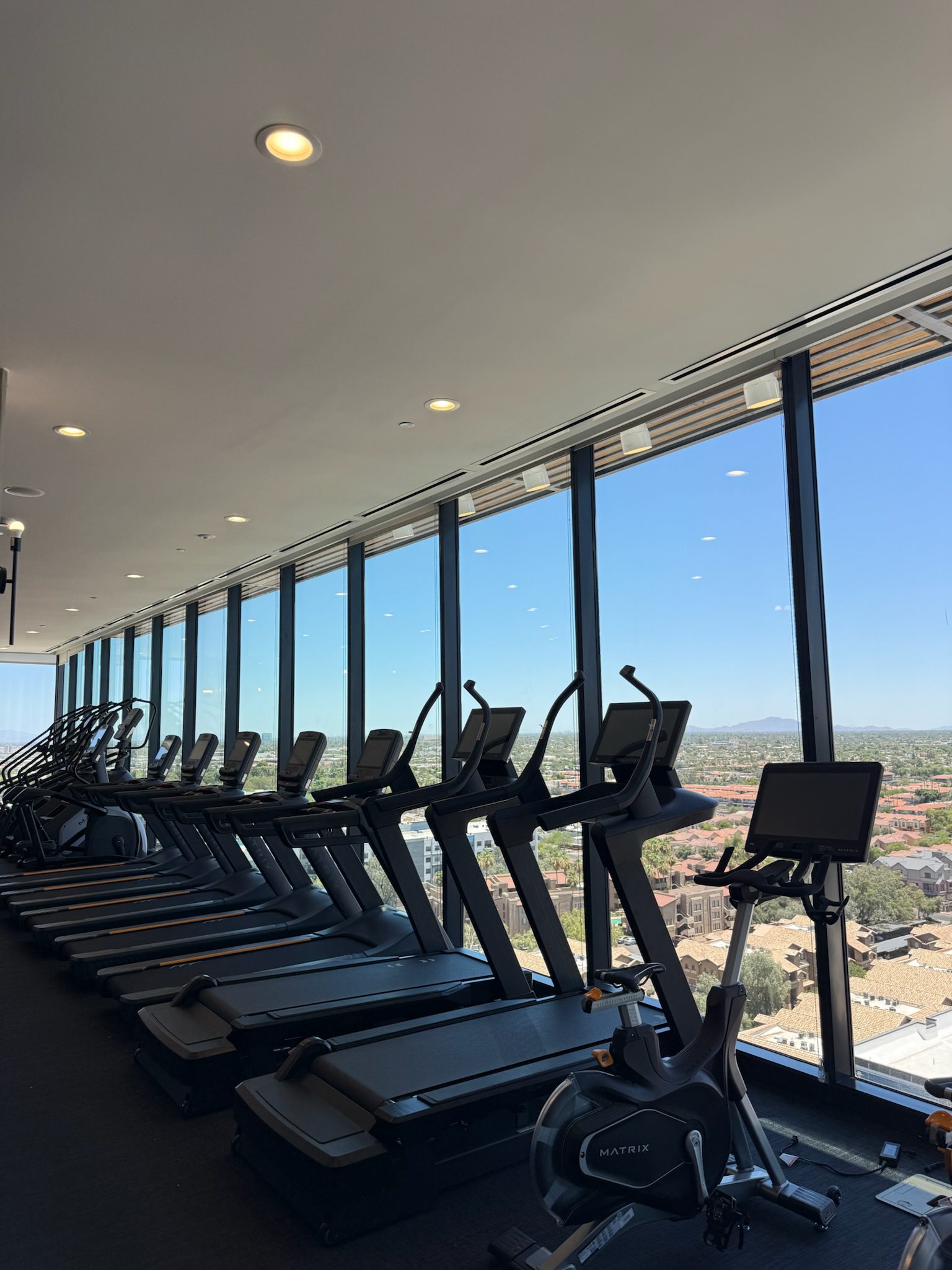 A row of treadmills and exercise bikes lines a gym with floor-to-ceiling windows, offering a panoramic view of a cityscape under a clear blue sky.