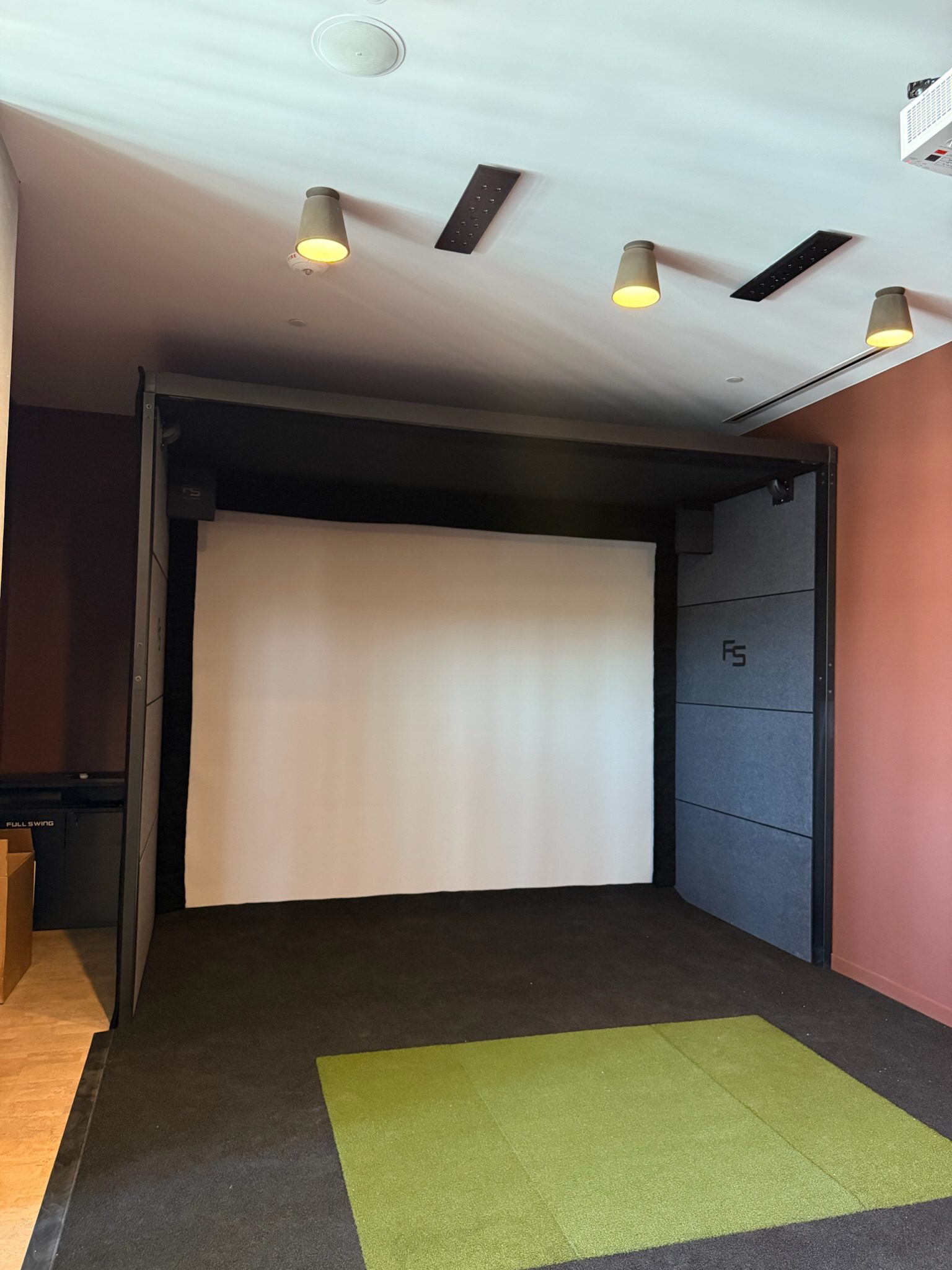 A golf simulator room with a white projection screen, overhead lights, black flooring, and a green rectangular turf mat in front of the screen. The walls are a mix of dark and light tones.