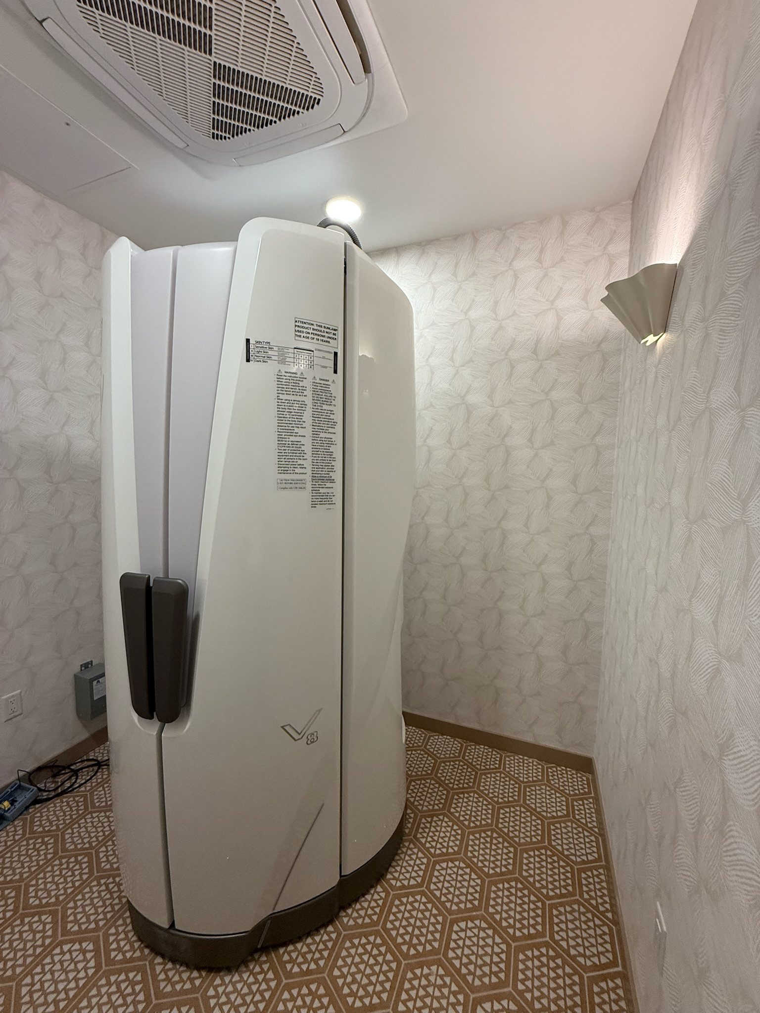 A large, white pod-like machine stands in a small room with patterned flooring and wallpaper. The machine has handles and a control panel, and is lit by ceiling and wall lights.