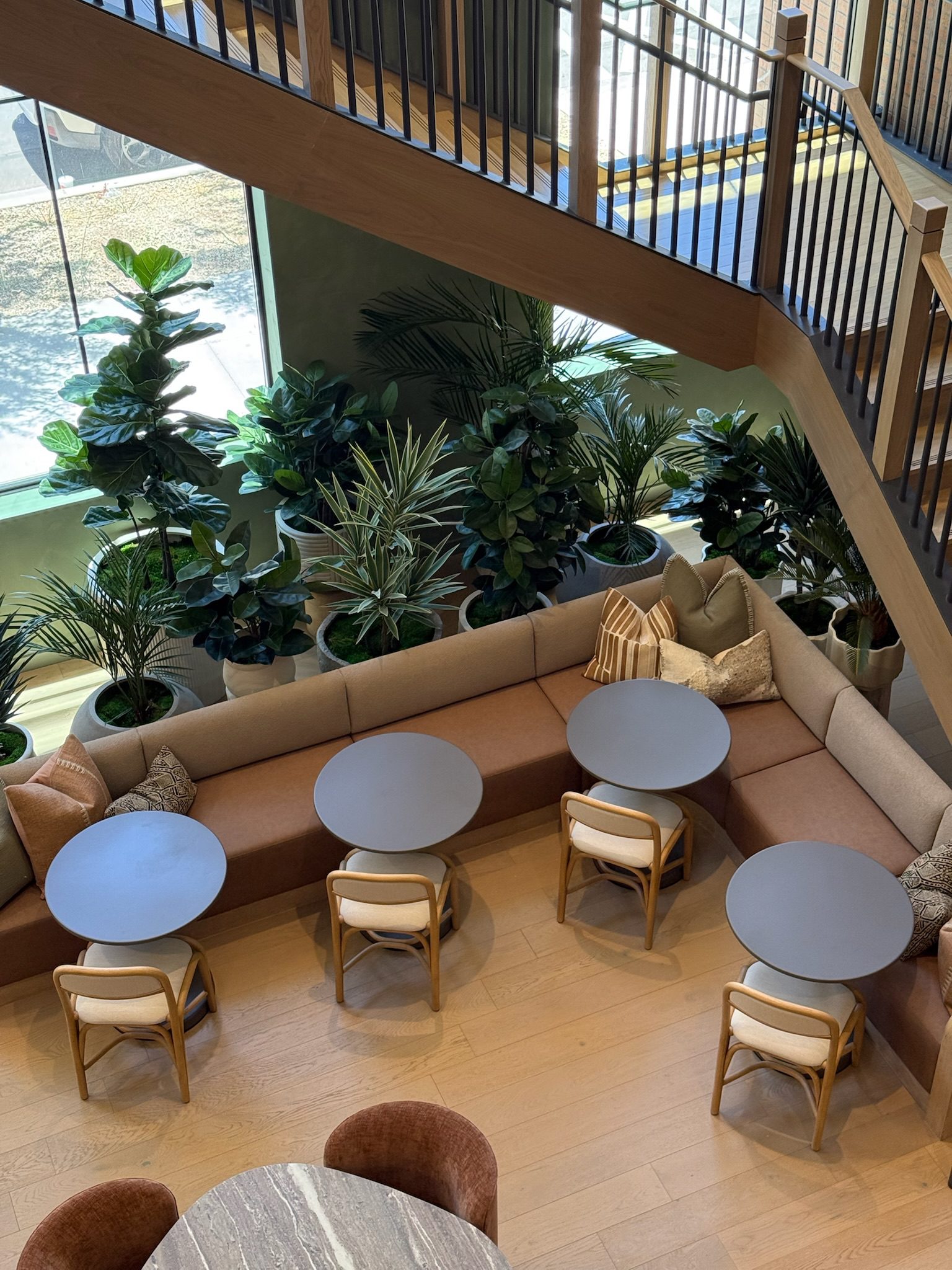 A modern cafe interior with round tables, light wooden chairs, a brown cushioned bench with pillows, and many green potted plants by a large window; stairs and railing are visible above.