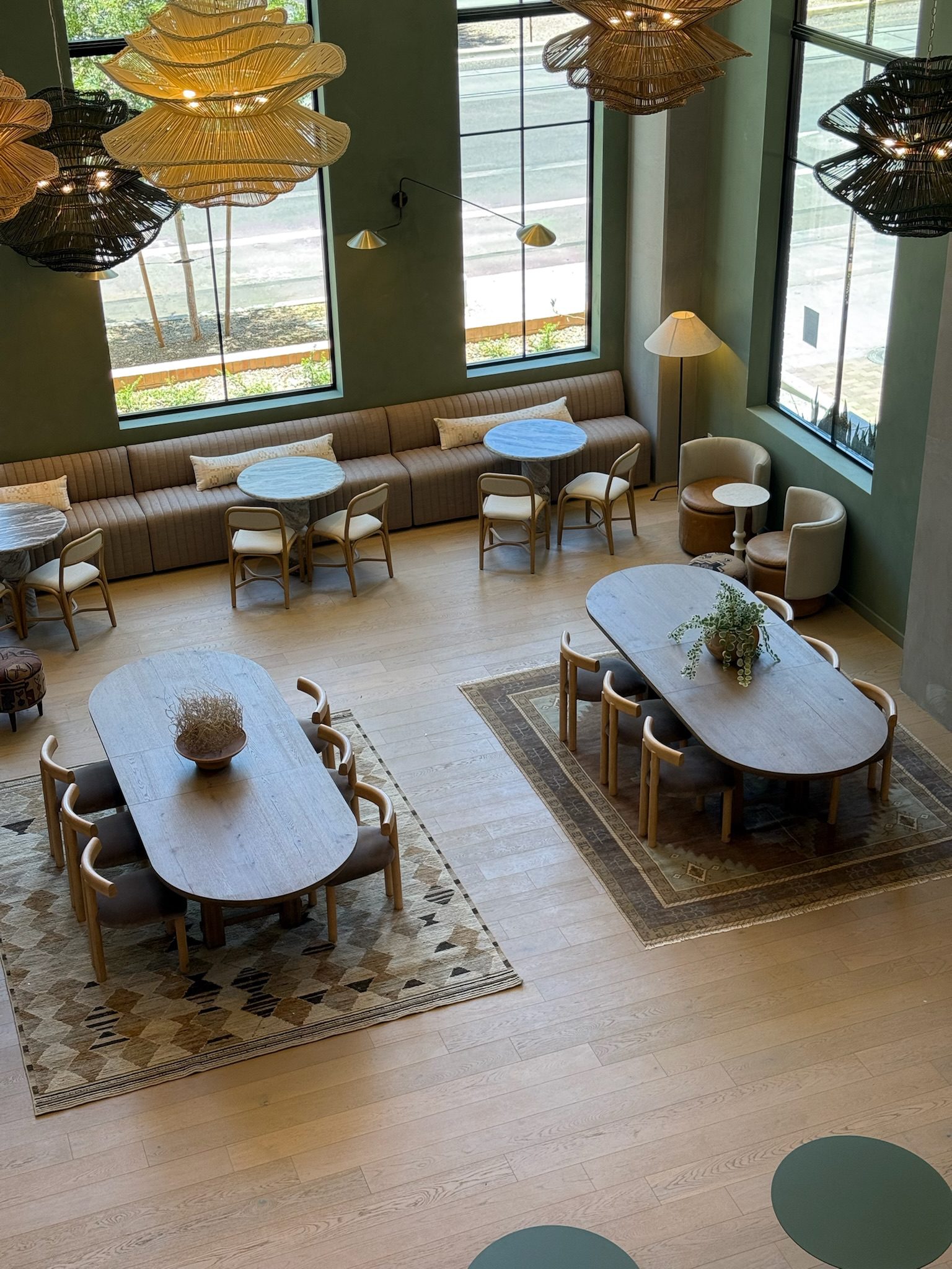 Modern café interior with large windows, oval wooden tables, chairs, patterned rugs, and a long cushioned bench against the wall. Woven pendant lights hang from the ceiling, and natural light fills the space.