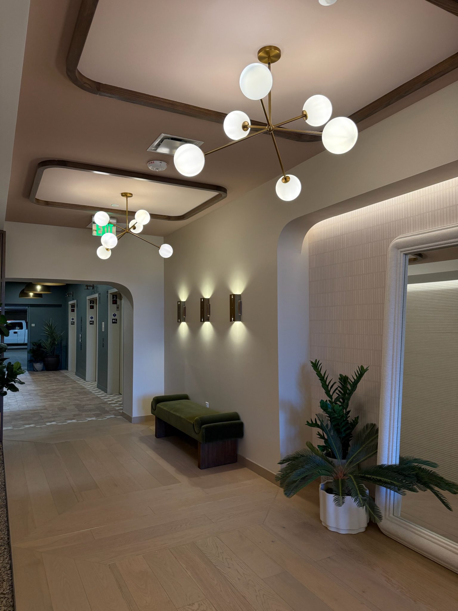 A modern hallway with light wood floors, spherical pendant lights, a green cushioned bench, potted plant, wall sconces, and a large mirror. Elevator doors are visible in the background.