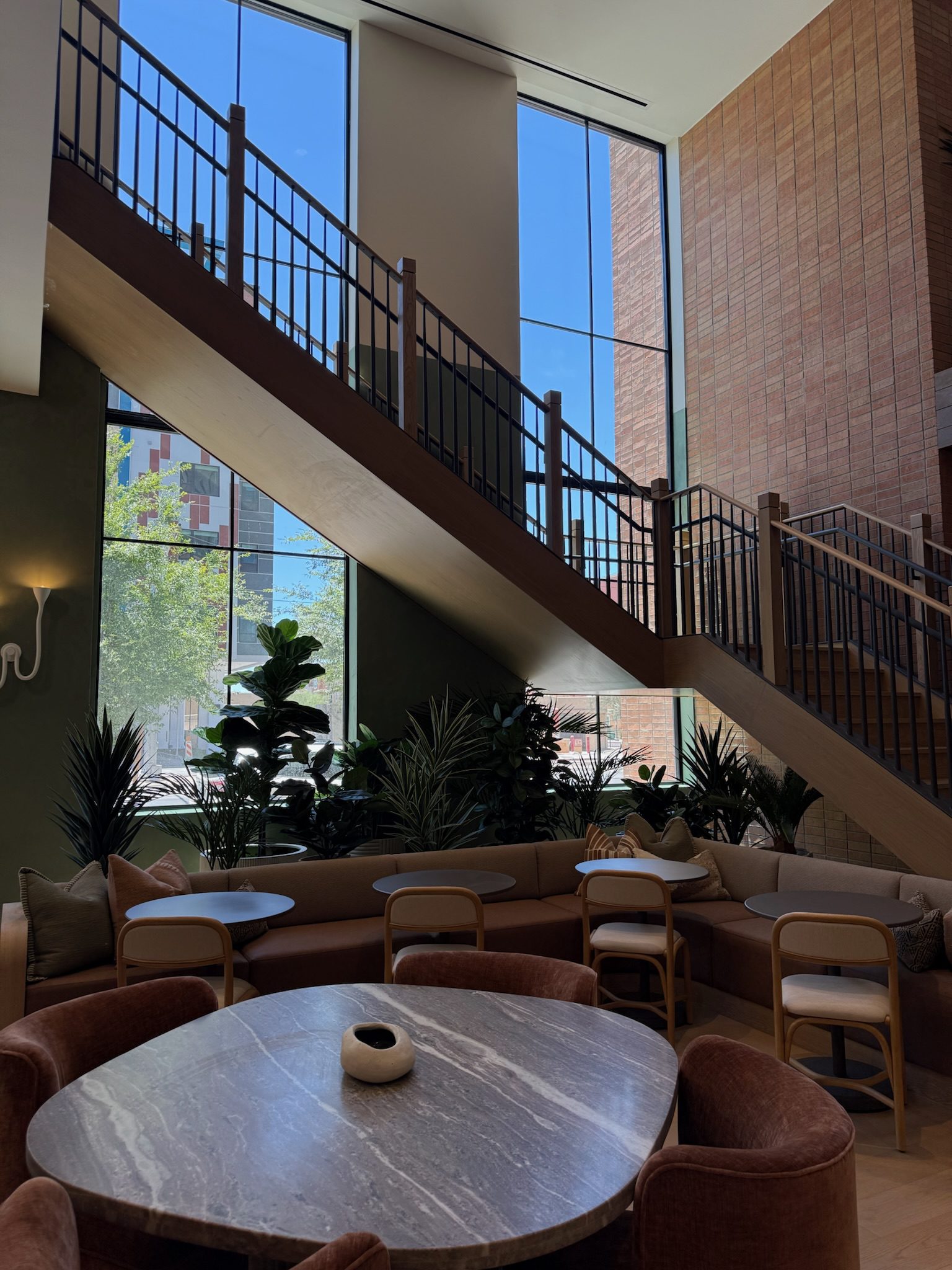 A modern lounge with round tables, brown chairs, cushioned benches, indoor plants, and large windows letting in natural light. A wooden staircase with a metal railing runs diagonally in the background.