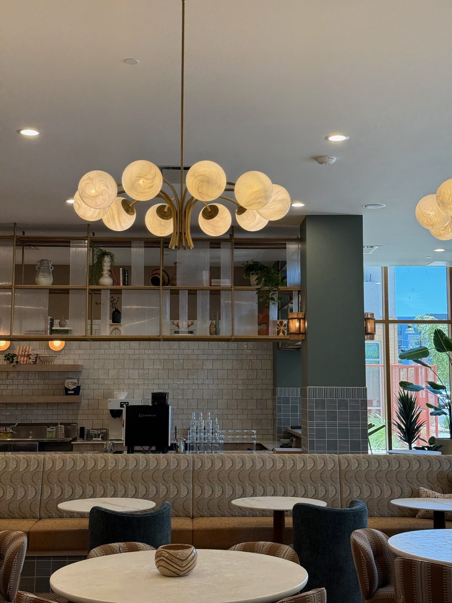 A modern cafe interior with round white tables, cushioned chairs, and a patterned banquette. Decorative light fixtures hang from the ceiling, and shelves with plants and pottery line the back wall above a tiled counter.