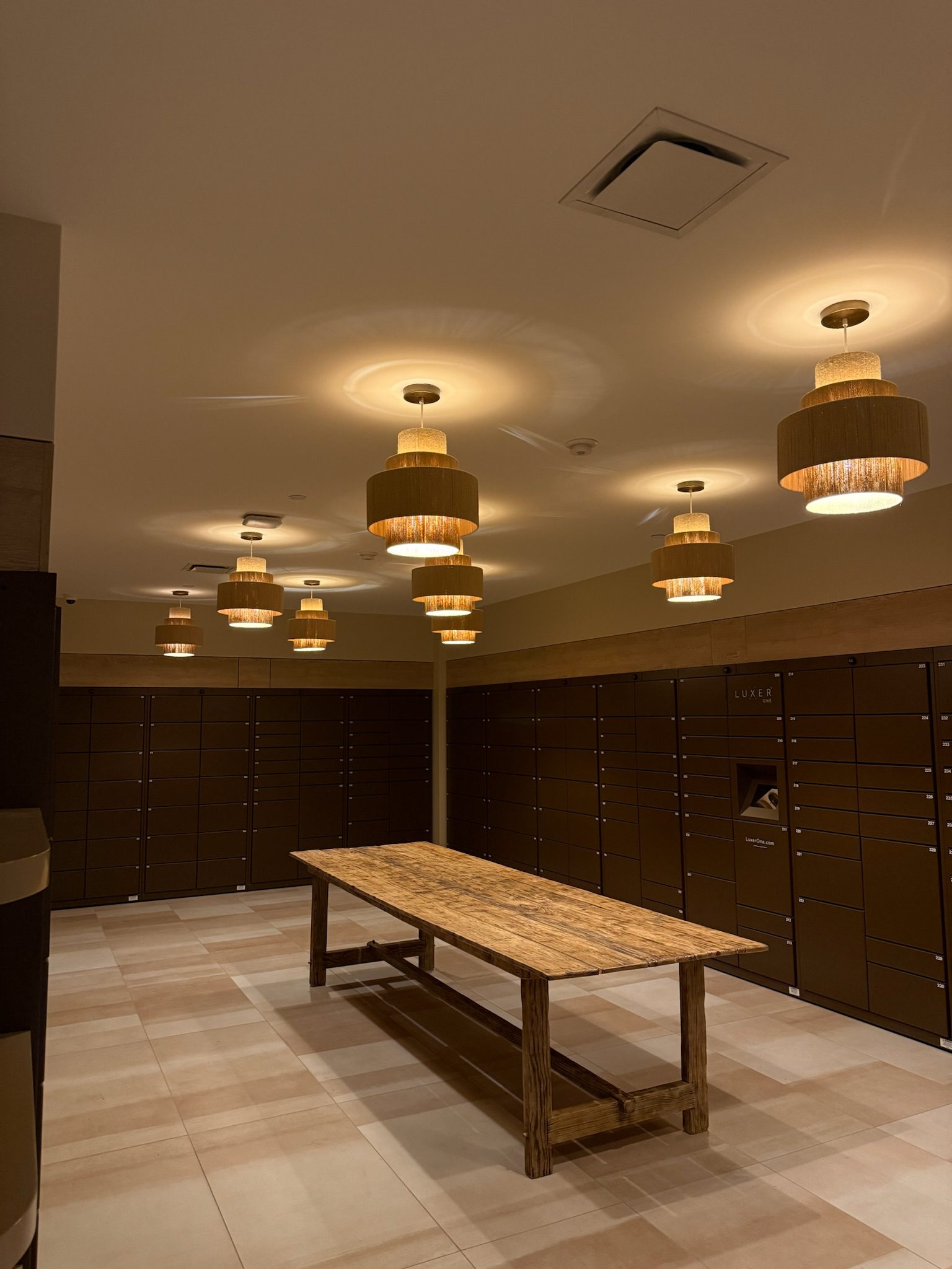 A spacious locker room with brown lockers lining the walls, a long wooden table in the center, and several modern pendant lights hanging from the ceiling, casting a warm glow.
