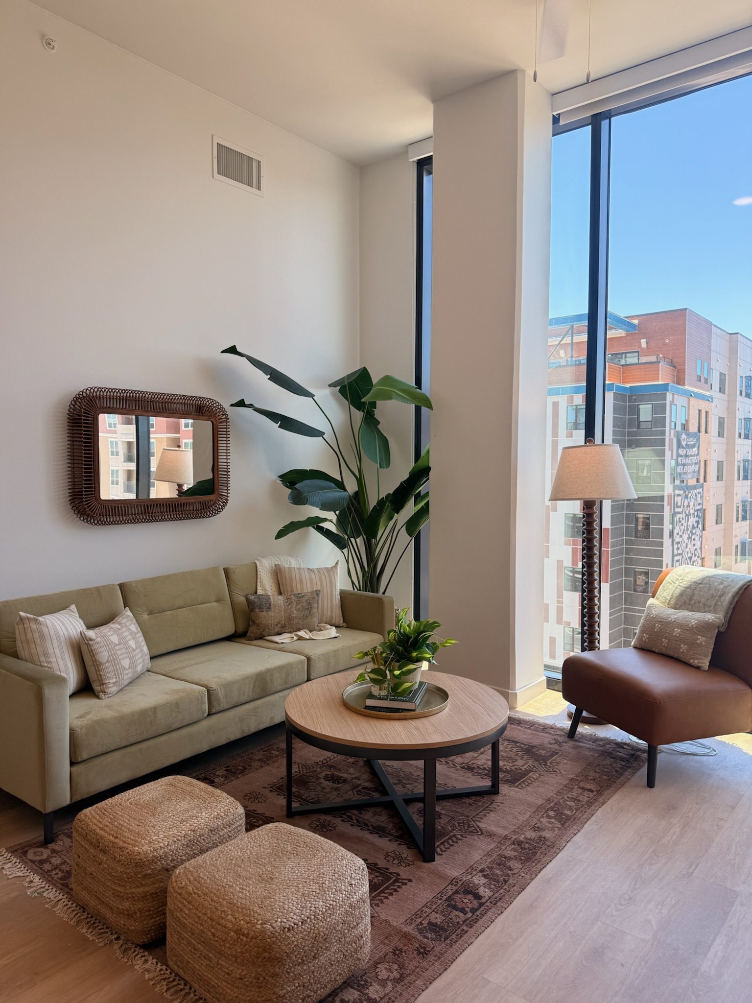 A modern living room with a green sofa, brown leather chair, two woven ottomans, a round wooden coffee table, potted plant, mirror, floor lamp, and large windows overlooking tall city buildings.