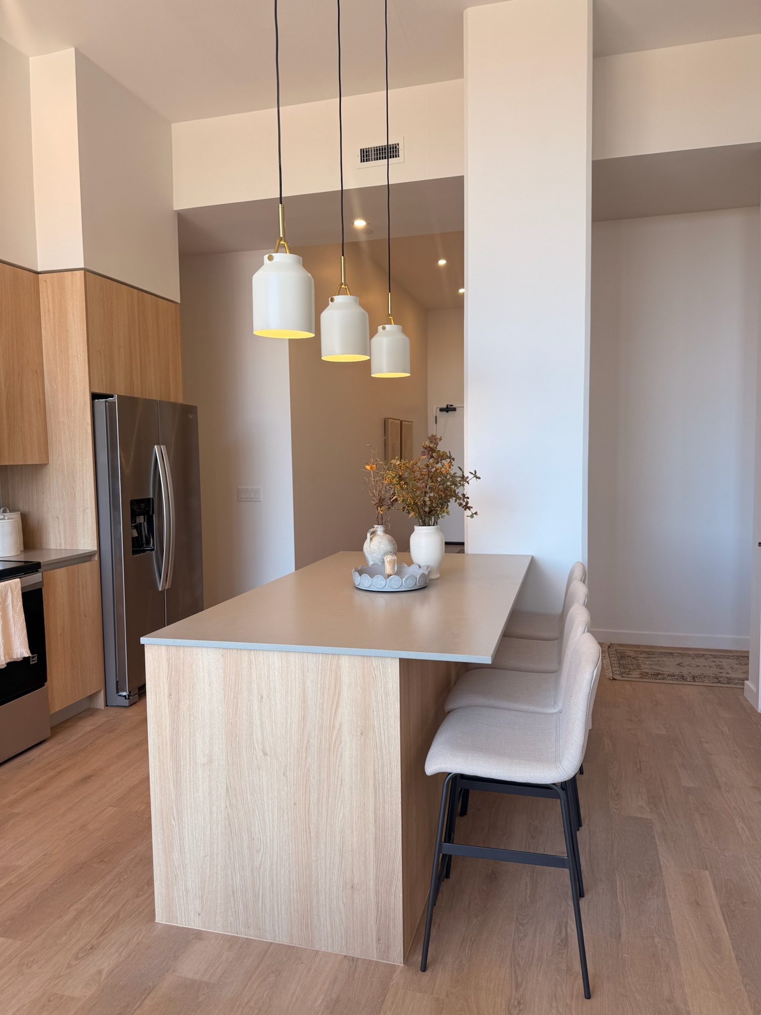 A modern kitchen with light wood cabinets, a gray countertop island, three beige cushioned bar stools, pendant lights, stainless steel appliances, and minimalist decor with a small vase and autumn branches.
