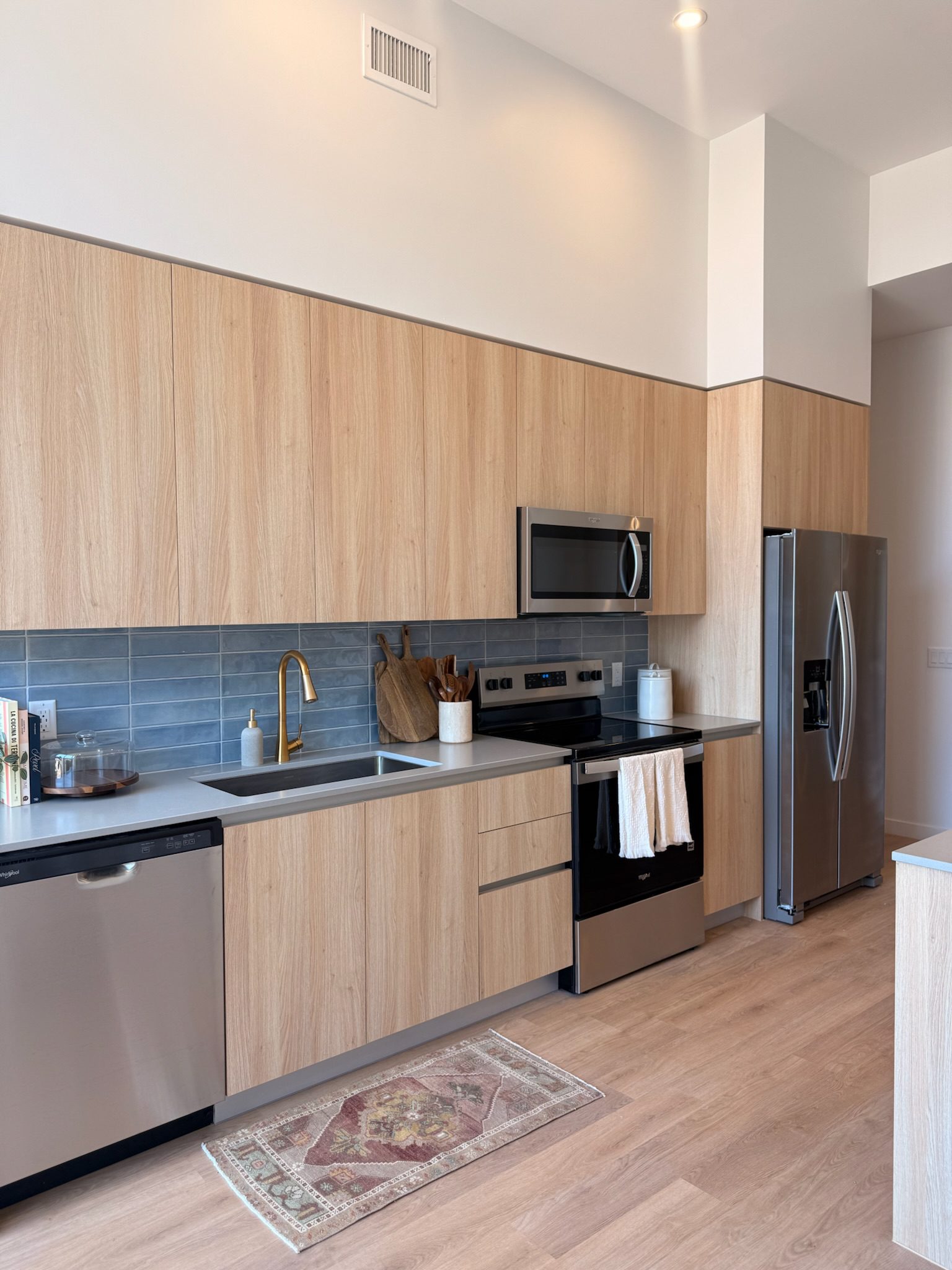 Modern kitchen with light wood cabinets, stainless steel appliances, gray tile backsplash, gold faucet, and a small patterned rug on light wood flooring. Countertops have decorative items and utensils.
