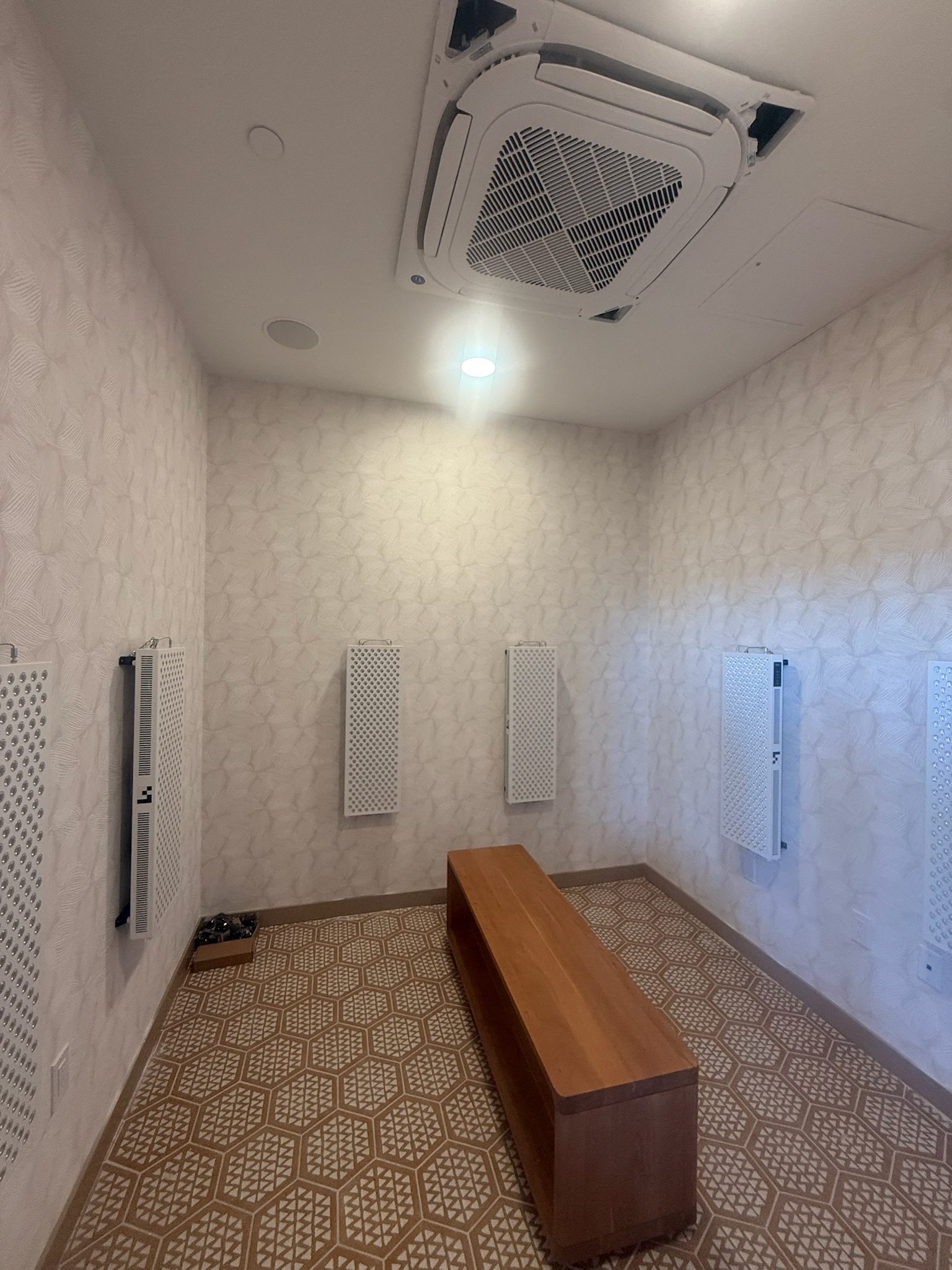 A small room with patterned walls and flooring, a wooden bench in the center, wall-mounted panels on three walls, and a ceiling air conditioning vent. The room is lit by a ceiling light.