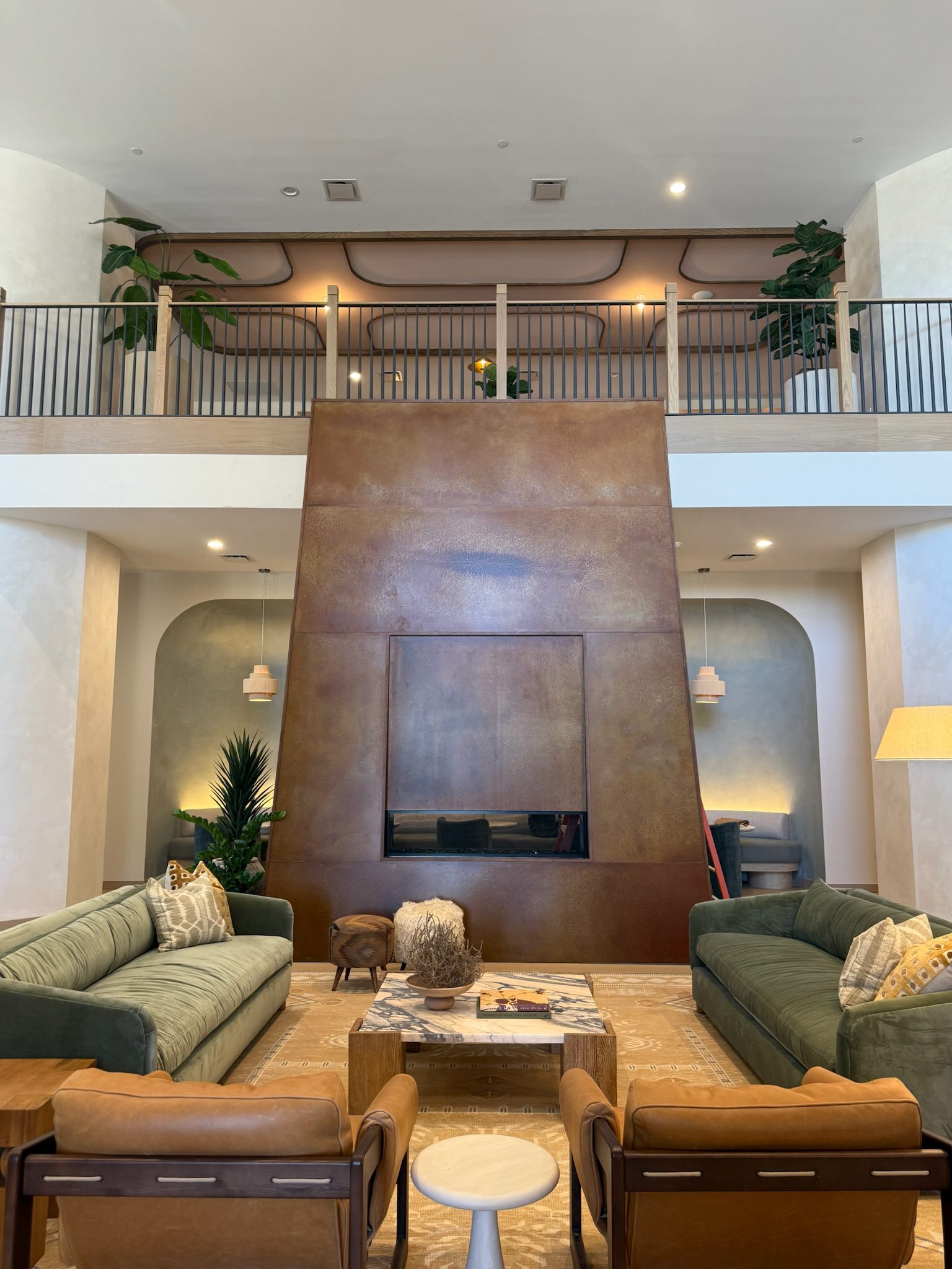 A modern, spacious living room with two green sofas, tan armchairs, a central coffee table, and a tall brown metal fireplace extending to the ceiling. An upper loft with railing overlooks the room.
