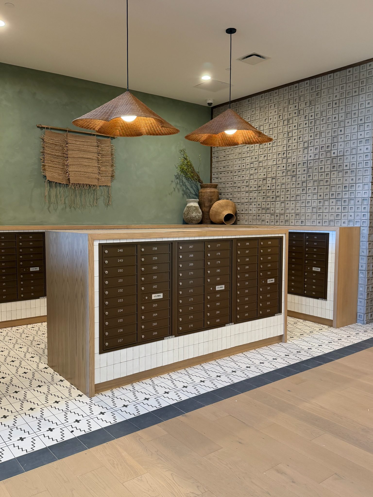 A modern mailroom with brown mailboxes set in a tiled counter, woven lampshades overhead, and ceramic vases and a woven wall hanging decorating the space. The floor features patterned tiles and light wood, offering a fresh construction update.