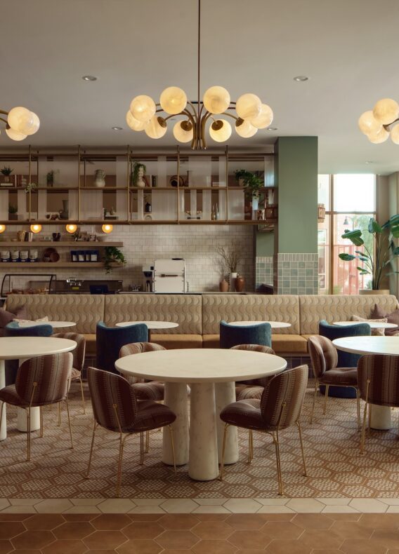 A modern cafe with round tables, cushioned chairs, pendant lights, a long booth, open shelving, potted plants, large windows, and a neutral color palette.